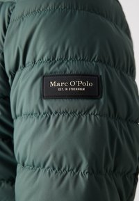 Roheline puffer-jakk, millel on õmblustega paneelid, sisaldab musta sildi, millel on kuldsete tähtedega kirjo "Marc O'Polo" ja "EST. IN STOCKHOLM."