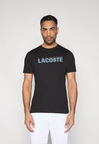Czarna bawełniana koszulka z niebieskim logo "LACOSTE". Posiada okrągły dekolt, krótkie rękawy, dopasowany krój oraz miękką teksturę.