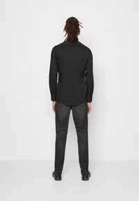 Črna majica z dolgimi rokavi z gladko teksturo in designom v slim fit, v kombinaciji s temno sivimi kavbojkami in črnimi čevlji, prikazana s hrbtne strani.