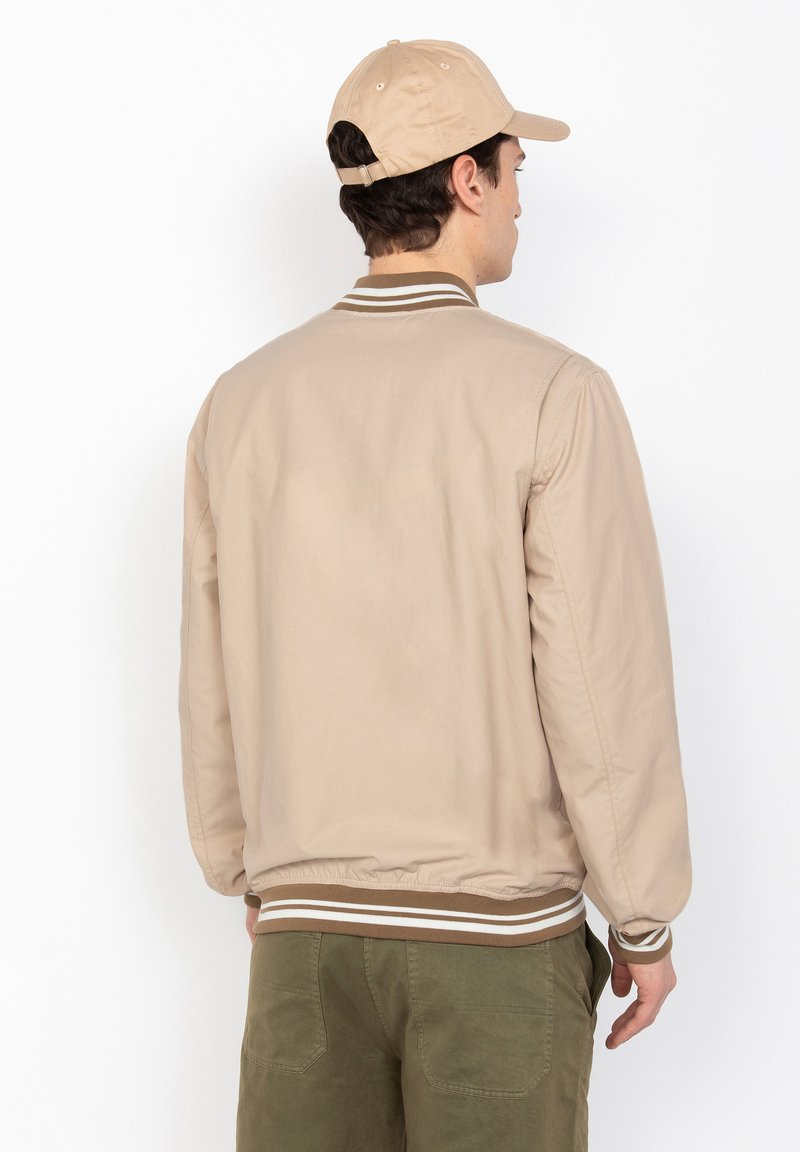 Beige Bomberjacke mit geripptem Kragen und Bündchen, die weiße und braune Streifen aufweist. Der Stoff wirkt glatt und leicht.