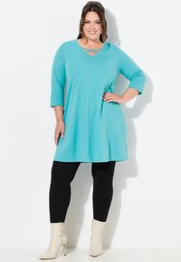 Turquoise tuniek met een V-hals en crisscross detail, 3/4 mouwen, gecombineerd met zwarte leggings en crèmekleurige enkellaarsjes. Gladde textuur.