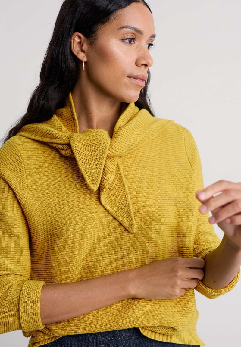 Mosterdgele geribbelde trui met een unieke knoopdetail bij de halslijn, voorzien van lange mouwen en een losse pasvorm.