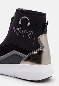 Czarne i metaliczno-srebrne wysokie trampki z białą podeszwą, z napisem "KARL" i grafiką słuchawek po boku.