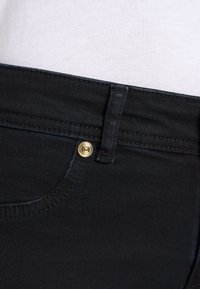 Black denim jeans feature a gold button and a stitched waistband. The fabric has a smooth texture with a slight sheen.