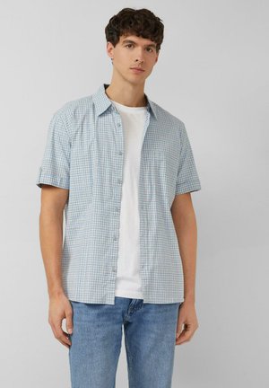 Jeune homme portant une chemise à manches courtes à carreaux bleu clair par-dessus un t-shirt blanc et un jean bleu, debout devant un fond uni.