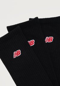 Τρία μαύρα κάλτσες πλήρωσης με κεντημένα κόκκινα και λευκά λογότυπα «NB» κοντά στην πάνω άκρη, τοποθετημένα επικαλυπτόμενα σε λευκό φόντο.