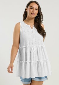 Vit ärmlös blus med rund halslinning, spetsbroderi och en tiers ruffle fåll. Inkluderar en dekorativ knut fram.