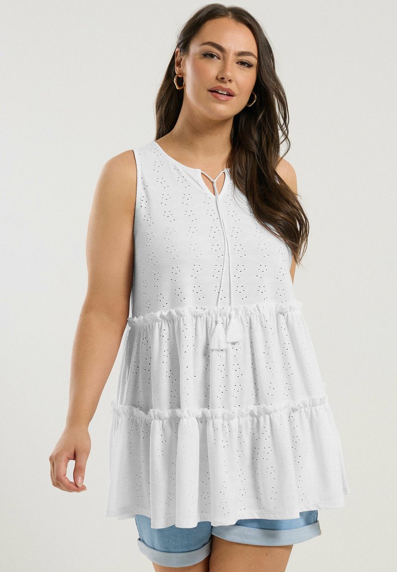 Vit ärmlös blus med rund halslinning, spetsbroderi och en tiers ruffle fåll. Inkluderar en dekorativ knut fram.