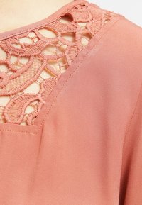 Blusa rosa con detalle de encaje en el escote, tela transparente y una textura suave, con un corte en forma de V debajo del encaje.