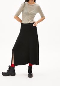 Šedý tričko s krátkým rukávem v kombinaci s černou maxi sukní s žebrovaným vzorem a bočním rozparkem, doplněné černými šněrovacími botami a červenými ponožkami.