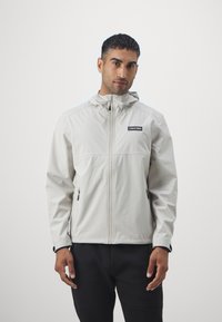 Giubbotto impermeabile grigio chiaro con chiusura a zip, cappuccio regolabile e due tasche laterali. Presenta una toppa con logo nera sul petto.