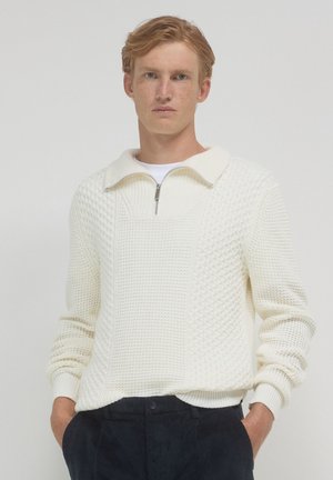 Maglione bianco lavorato a maglia con colletto a zip quarti, motivo testurizzato, polsini a coste e vestibilità rilassata. Indossato sopra una camicia bianca.