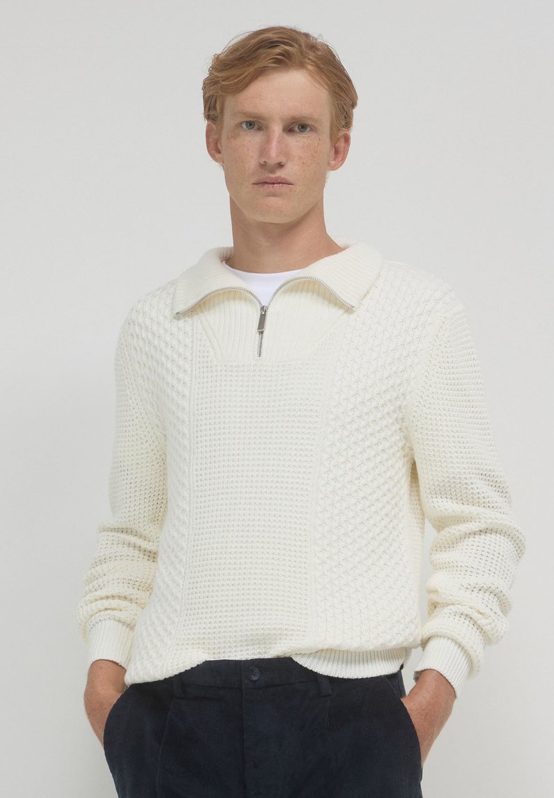 Maglione bianco lavorato a maglia con colletto a zip quarti, motivo testurizzato, polsini a coste e vestibilità rilassata. Indossato sopra una camicia bianca.