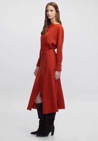 Vestido rojo de manga larga con una abertura alta y cintura con cordón. Cuenta con una tela suave y detalles de botones en las mangas. Se lleva con botas marrones hasta la rodilla.