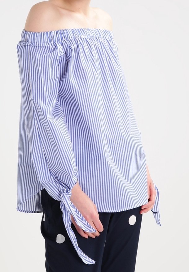 Blusa de rayas azul y blanca, con escote off-the-shoulder, cuello fruncido y mangas atadas, combinada con pantalones azul marino con lunares blancos.