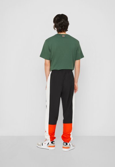 T-shirt en coton vert associé à un pantalon de jogging noir et orange avec des bandes blanches sur les côtés, des poignets élastiques et des baskets sportives.