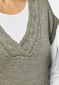 Grau gestrickte Weste mit V-Ausschnitt und strukturierter Zopfmusterung. Getragen über einem weißen Langarmoberteil, das ein weiches, leichtes Material zeigt.