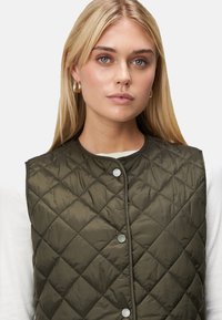 Gequilted olijfgroen vest met een ronde halslijn, drukknopen en een soepele textuur, gelaagd over een wit shirt.