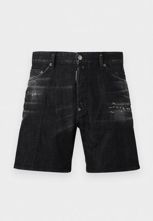 Young man standing with one hand in pocket, wearing a checkered button-up shirt, white tank top, black denim shorts, white socks, and black sneakers.