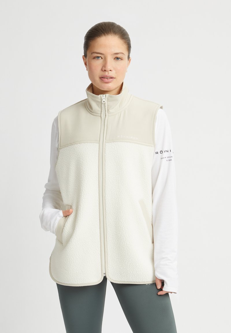 Kvinna klädd i en krämfärgad zip-up fleece-väst, vit långärmad undertröja och mörkgrå leggings, stående mot en enkel vit bakgrund.