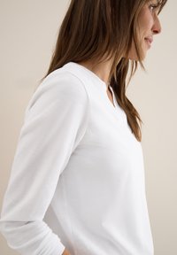 Weißes Langarmoberteil mit Henley-Ausschnitt, weicher Textur, schmaler Schnitt und dezenten Nähten an den Seiten.