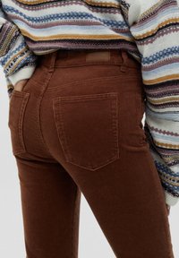 Bruine corduroy-broeken met een getailleerd ontwerp, voorzien van achterzakken en een stof met een geribbelde structuur. Gestyled met een gestreepte trui erboven.