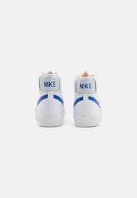 Bílé vysoké tenisky s modrými akcenty, se strukturovaným povrchem a logem "NIKE" na zadní straně. Gumové podrážky.