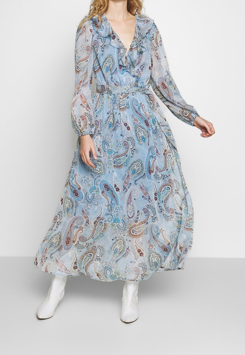 Femme portant une longue robe bleu clair à motif paisley avec un col à volants et des manches longues transparentes, associée à des bottines blanches.