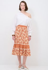 Weißes Oberteil mit einseitiger Schulterpartie, kombiniert mit einem langen, orangefarbenen Rock im floralen Muster mit einem gestuften Saum und karierter Borte. Weiße Schuhe.