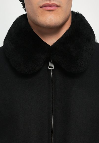 Schwarzer Mantel mit einem luxuriösen schwarzen Pelzkragen, einem mit Reißverschluss versehenen Vorderteil und einer glatten Stoffstruktur, die sich auf minimalistische Designelemente konzentriert.