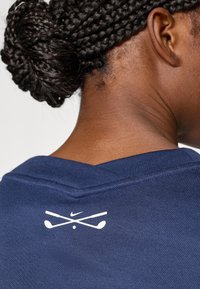 Μπλε ναυτικό πόλο πουκάμισο με υφασμάτινη υφή· διαθέτει λευκό λογότυπο Nike και διασταυρωμένα γκολφ ρόπαλα στην πλάτη.