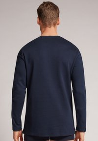 Námořnická modrá košile s dlouhým rukávem z měkkého materiálu, s kulatým výstřihem a rovně střiženým lemováním. Minimalistický design bez viditelných vzorů nebo akcentů.