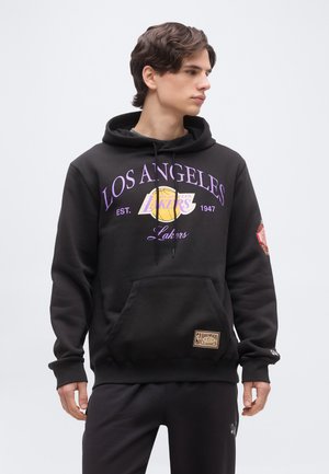 Jeune homme portant un sweat à capuche noir des Los Angeles Lakers avec un texte violet et un logo de basketball jaune, debout devant un fond clair uni.
