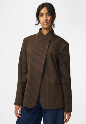 Jeune femme aux cheveux foncés porte un blazer marron foncé à fines rayures avec un col asymétrique boutonné et un jean bleu.