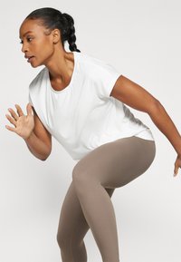 T-shirt de sport blanc à manches courtes associé à des leggings beiges moulants. Le tissu semble lisse et extensible, adapté au mouvement et à l'exercice.