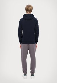 Navy kék kapucnis felső, laza fazonnal, kenguru zsebbel és fehér logóval a kézen. Szürke melegítőnadrággal és fehér tornacipővel párosítva.