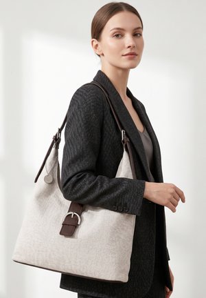Vrouw in een donker colbert die een grote beige draagtas met een bruine riem en gesp over de schouder draagt tegen een witte achtergrond.
