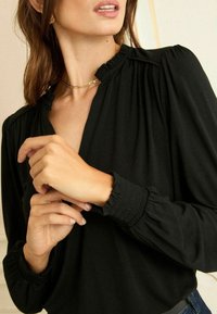Fekete blúz magas nyakkivágással, ráncolt vállakkal és gumírozott mandzsettákkal. Simasága mellett finom ráncolás jellemzi a gombolás mentén.