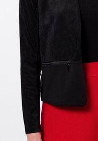 Veste noire avec une finition texturée et douce, à manches longues, dotée d'une poche zippée sur le bas à l'avant, associée à une jupe rouge.