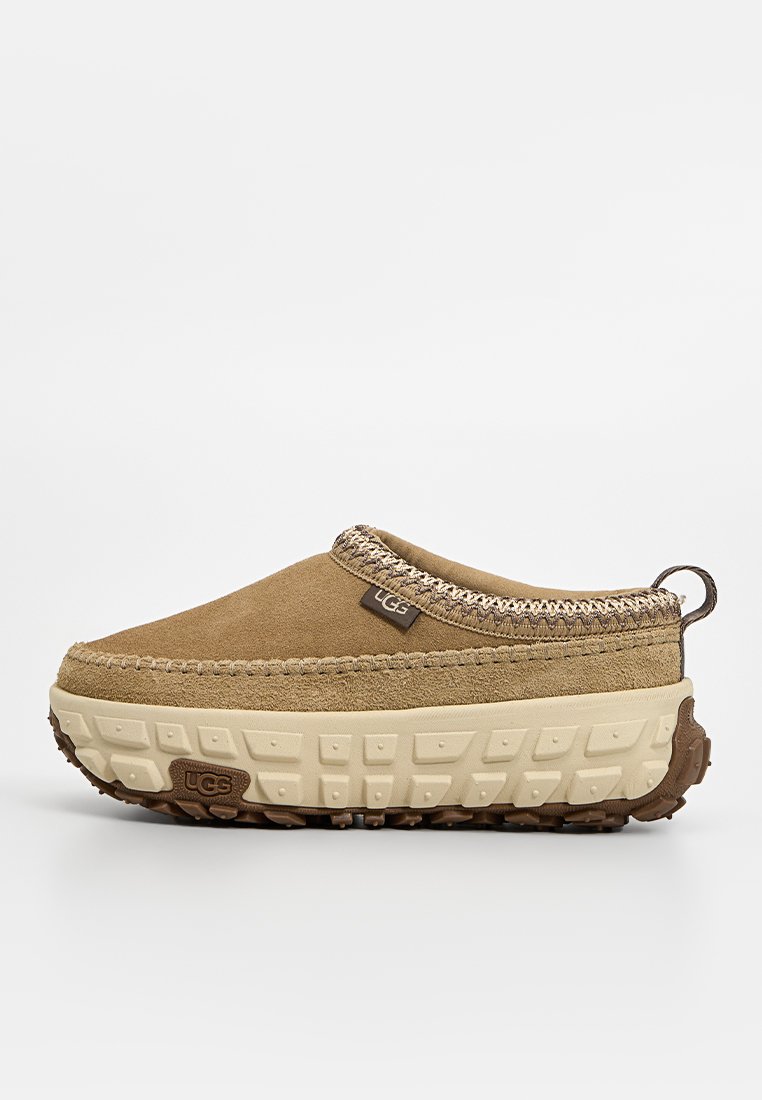 Zapato slip-on de ante color tostado con acentos texturizados en marrón, que presenta una suela robusta de color crema y detalles trenzados a lo largo del cuello. Logotipo de UGG presente.