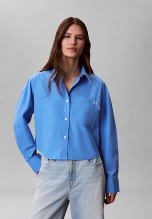 Ung kvinne med langt brunt hår har på seg en knallblå cropped skjorte med knapper og lyseblå høytlivsjeans mot en ensfarget grå bakgrunn.