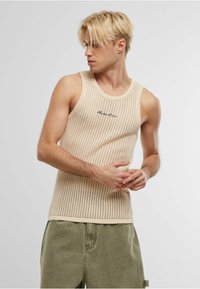 Beige strikket tanktop med åpen rutenettmønster, rund hals og subtil svart brodert tekst, kombinert med olivengrønne shorts.