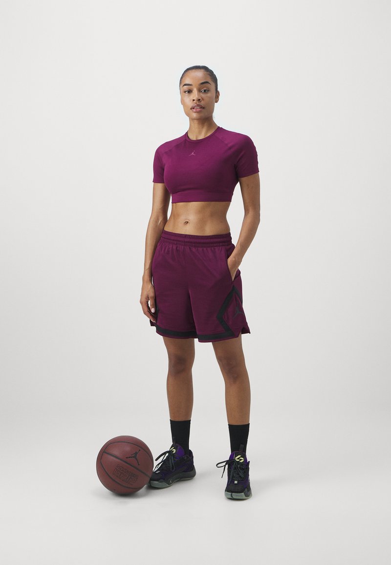 Maroon verkürzter Sporttop und passende Shorts mit schwarzen Akzenten. Schwarze Basketballschuhe und ein Basketball liegen neben ihr auf dem Boden.