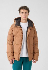 Giacca imbottita marrone con cappuccio, caratterizzata da un design bicolore. Realizzata in materiale liscio e imbottito con chiusura a cerniera, indossata sopra una camicia bianca.