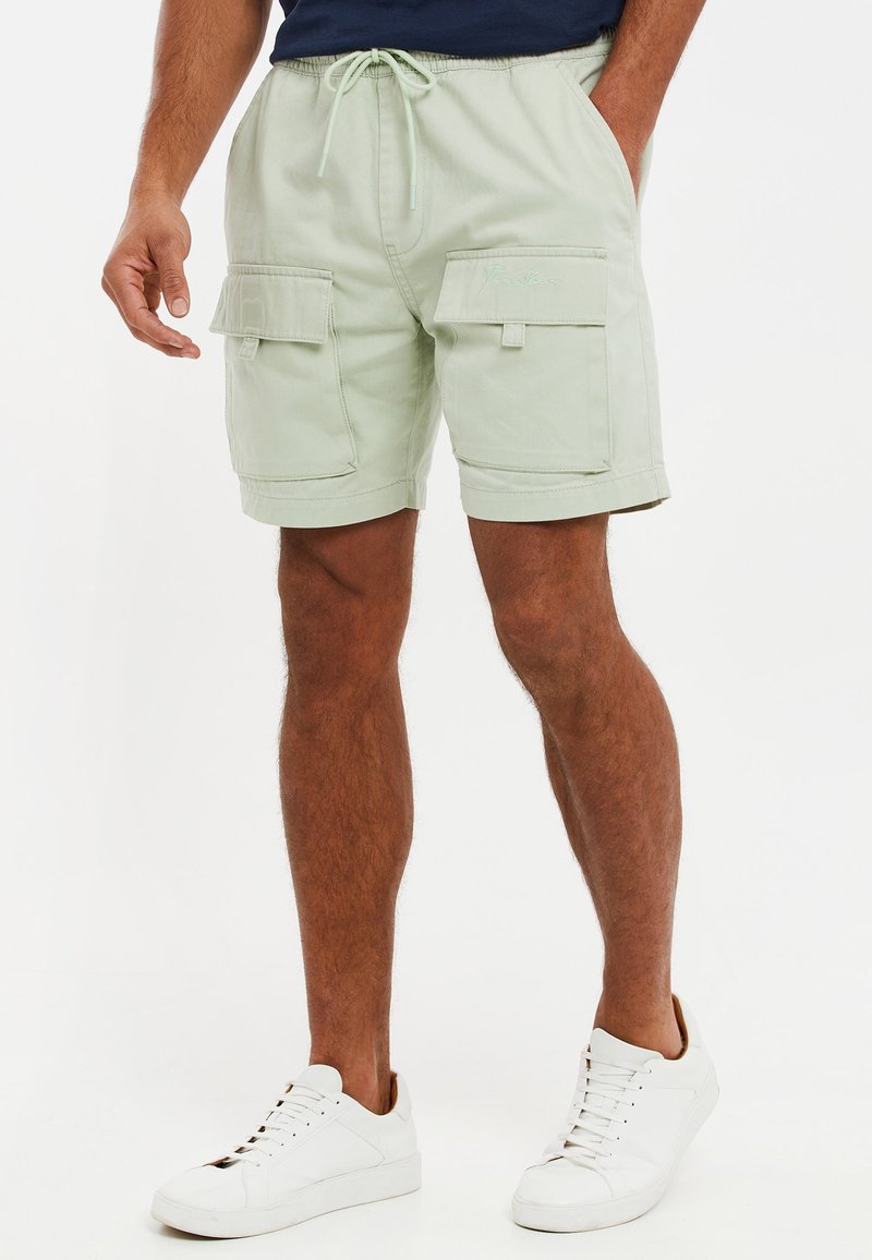 Homme portant un short cargo vert clair avec taille à cordon et des baskets blanches, debout, les mains partiellement dans les poches.