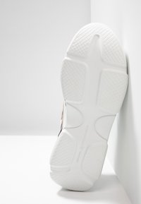Semelle blanche de basket avec motifs de grip texturés et marquage "KENNEL & SCHMENGER", positionnée verticalement contre un mur blanc.
