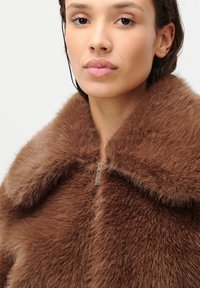 Καφέ ψεύτικο γούνινο παλτό με φαρδύ γιακά και κλείσιμο με φερμουάρ. Διαθέτει απαλή, υφασμάτινη επιφάνεια με πλούσιο, ζεστό χρώμα.