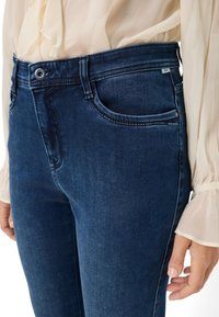 Jeans in denim di un blu scuro con una texture liscia, dotati di una tasca frontale, passanti per cintura e un dettaglio in metallo sul cinturino.