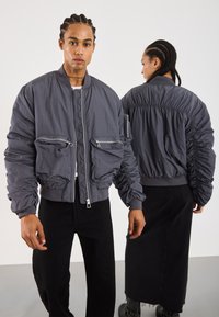 Twee mensen met gevlochten haar die bijpassende oversized grijze bomberjacks dragen, waarbij de een naar voren kijkt en de ander naar achteren.