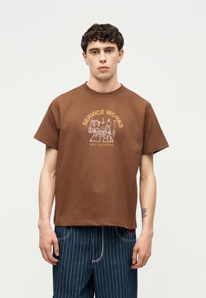 Νέος με κοντά σγουρά μαλλιά, φοράει καφέ μπλουζάκι με το κείμενο και το γραφικό «Service Works Soup for the Soul», συνδυασμένο με μπλε παντελόνια με ρίγες.